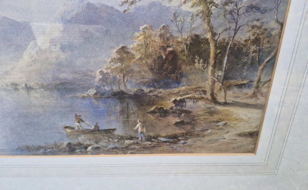 A framed 19th-century watercolour painting depicting a Lake District scene with small boats on a lake surrounded by trees and mountains, enclosed in a cream wash mount and a gilded decorative frame. View X