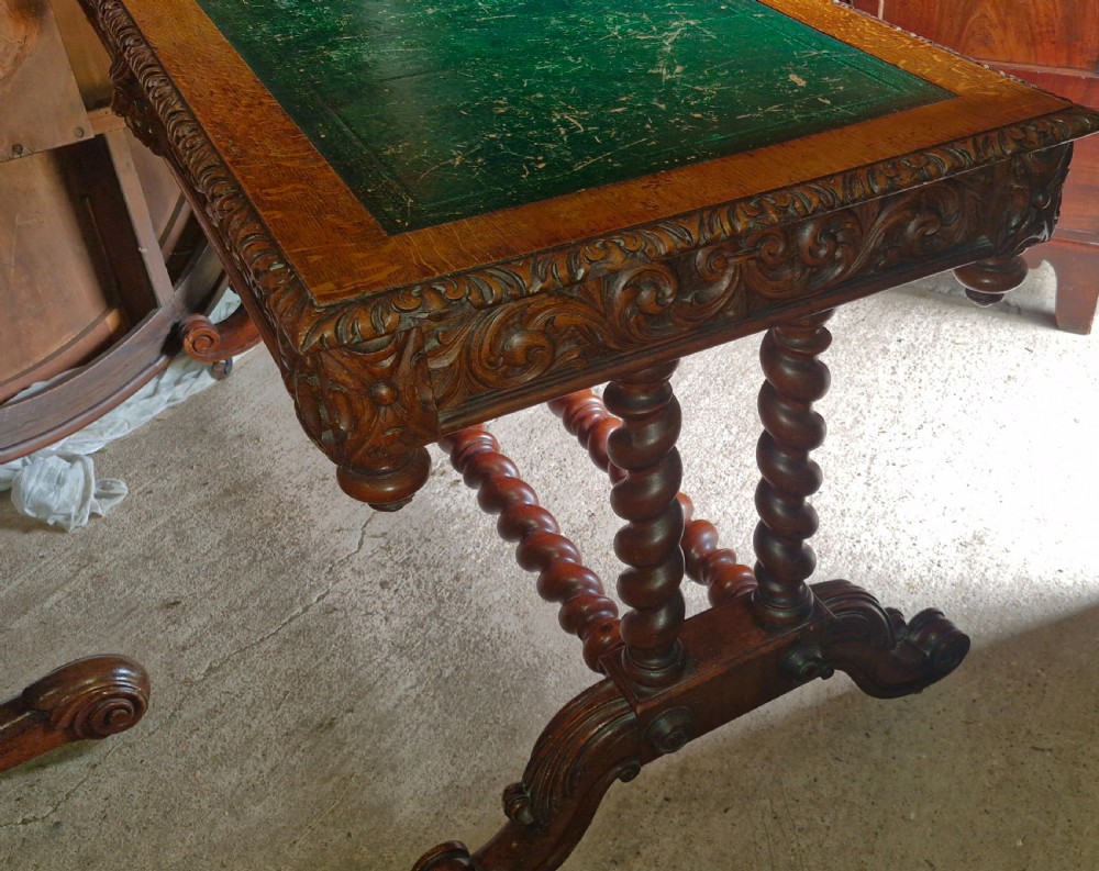 A Victorian carved oak writing desk featuring a green leather inset top, detailed Green Man mask carvings on the drawers, and turned barley twist stretchers. View VI