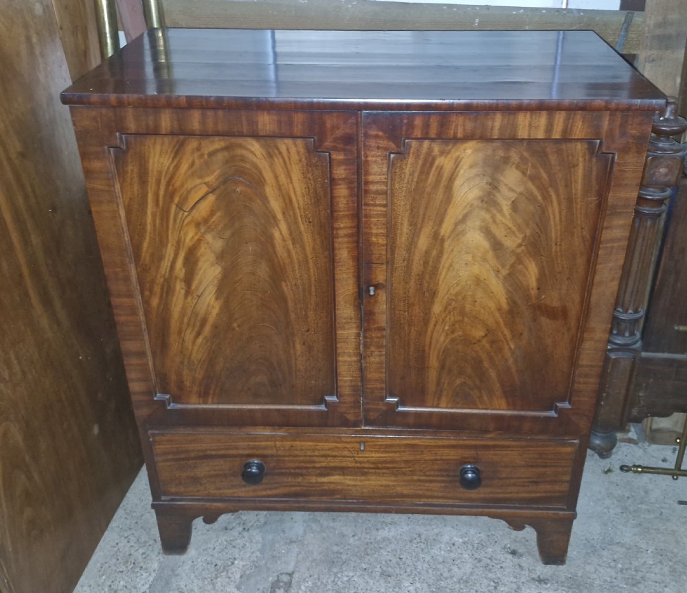 An early 19th-century mahogany dwarf linen cupboard featuring flame mahogany door panels, a single bottom drawer with turned knobs, and shaped bracket feet.