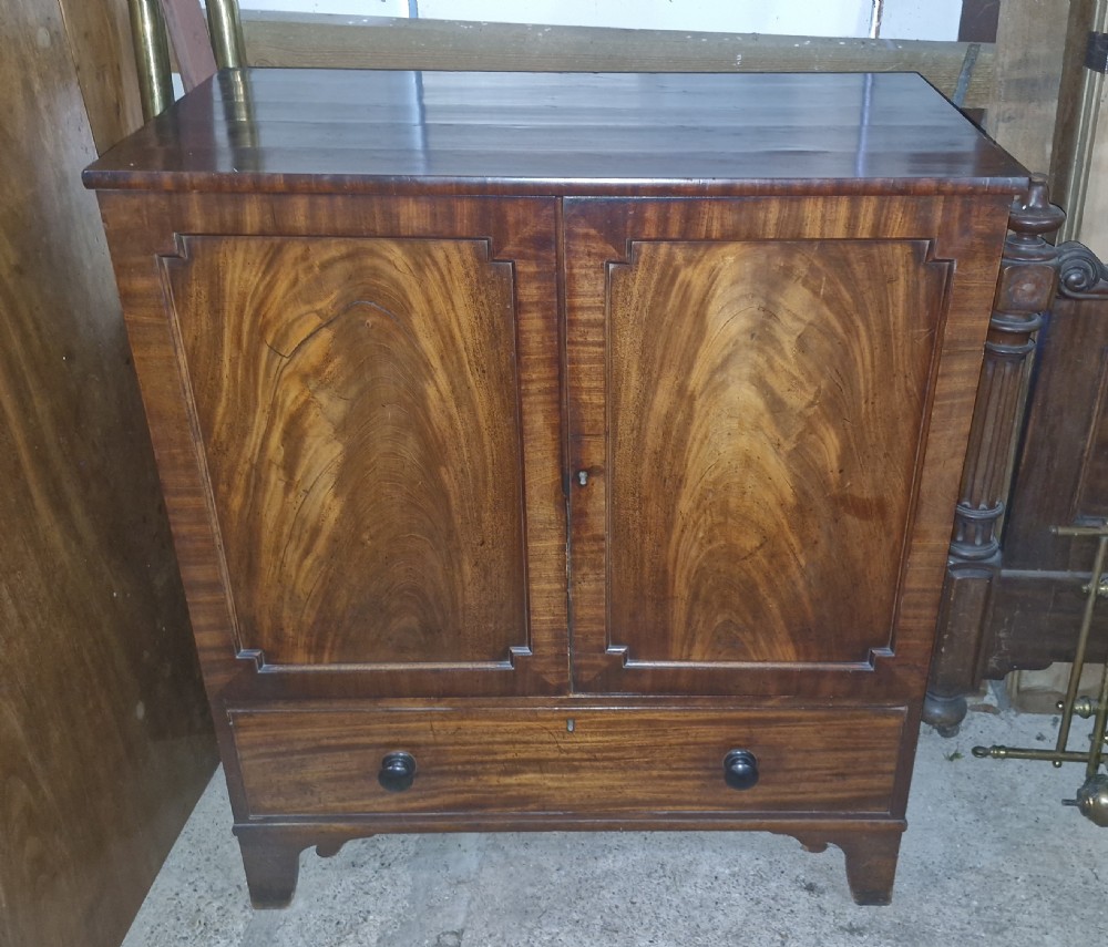 An early 19th-century mahogany dwarf linen cupboard featuring flame mahogany door panels, a single bottom drawer with turned knobs, and shaped bracket feet. View VI