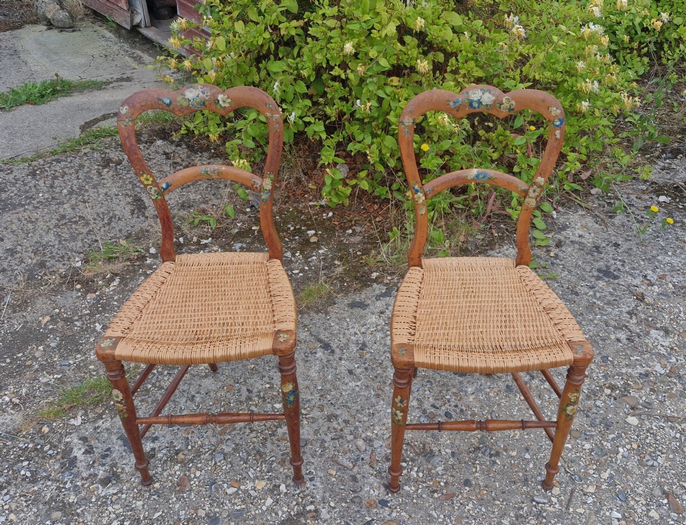 A pair of Victorian side chairs featuring light brown wooden frames adorned with hand-painted floral motifs and woven cane seats.