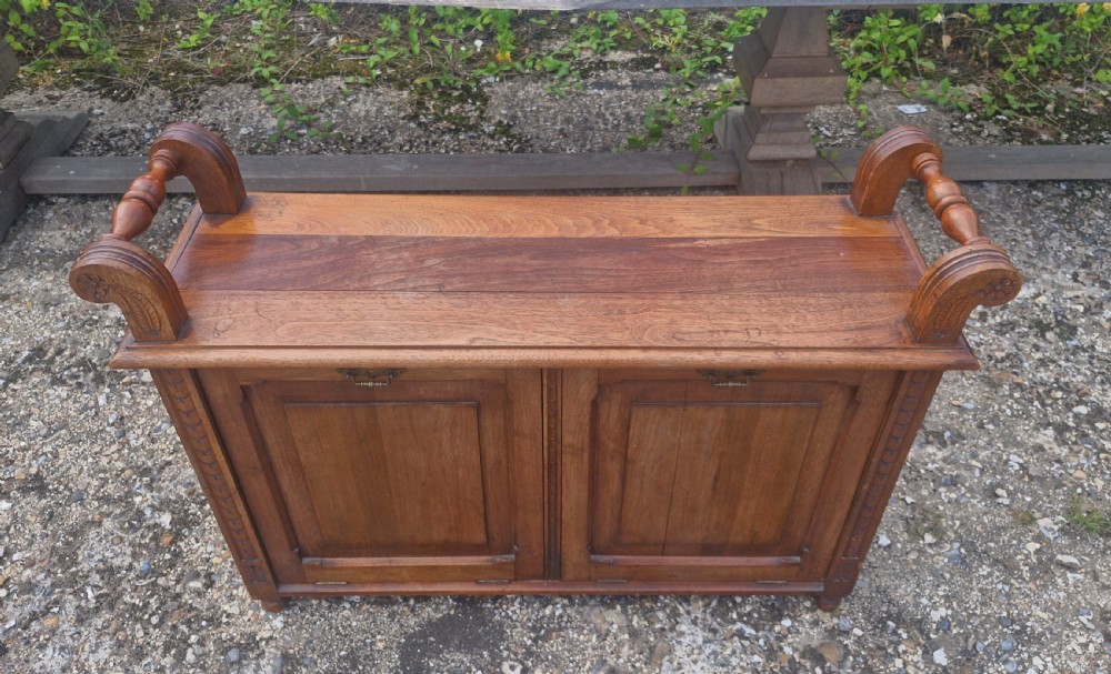 A late Victorian carved oak duet stool with two panelled cabinet doors, brass drop handles, and raised side scroll handles. View X