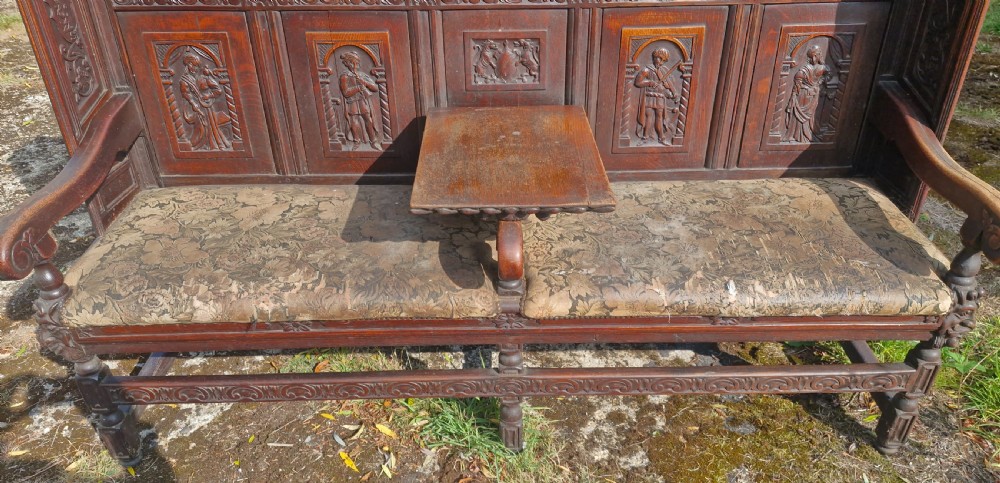 A dark carved oak hall bench featuring relief-carved figure panels on the high back, padded upholstered seats, and a central drop-down table section. View III