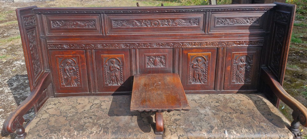 A dark carved oak hall bench featuring relief-carved figure panels on the high back, padded upholstered seats, and a central drop-down table section. View V