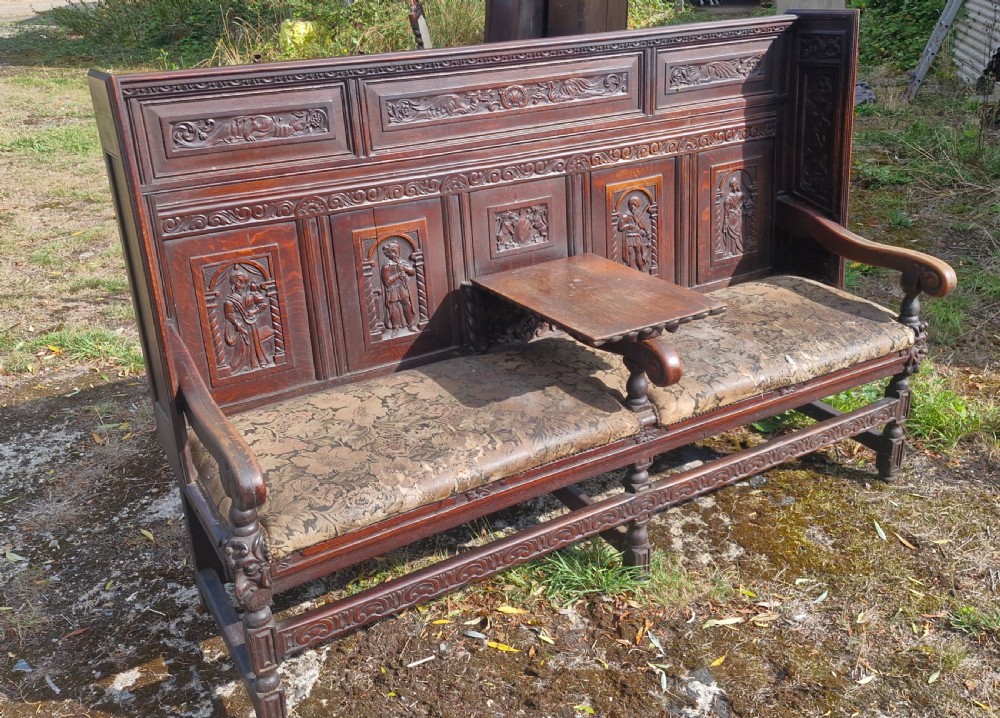 A dark carved oak hall bench featuring relief-carved figure panels on the high back, padded upholstered seats, and a central drop-down table section. View VI