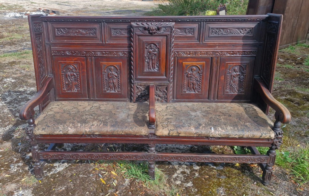 A dark carved oak hall bench featuring relief-carved figure panels on the high back, padded upholstered seats, and a central drop-down table section. View VII