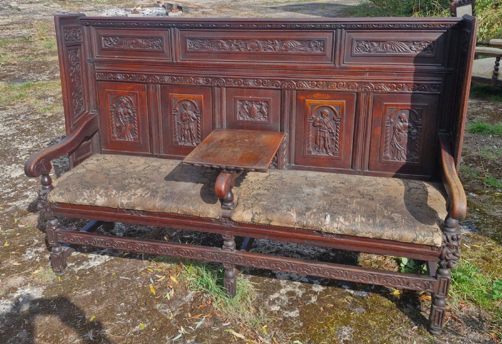 A dark carved oak hall bench featuring relief-carved figure panels on the high back, padded upholstered seats, and a central drop-down table section. View VIII