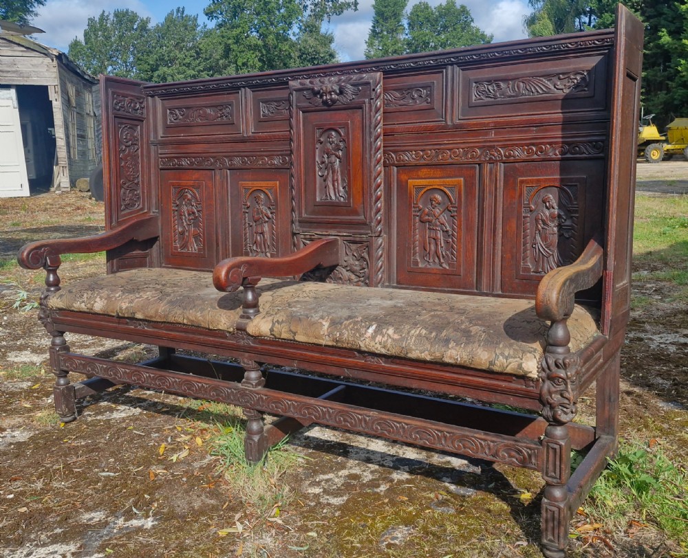A dark carved oak hall bench featuring relief-carved figure panels on the high back, padded upholstered seats, and a central drop-down table section. View IX