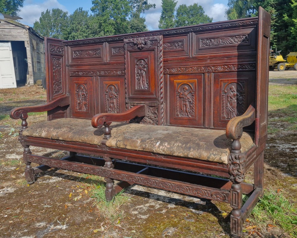 A dark carved oak hall bench featuring relief-carved figure panels on the high back, padded upholstered seats, and a central drop-down table section. View X