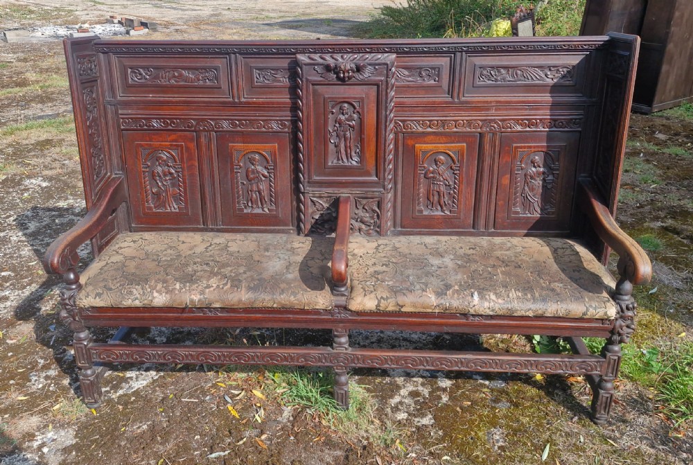 A dark carved oak hall bench featuring relief-carved figure panels on the high back, padded upholstered seats, and a central drop-down table section. View XI