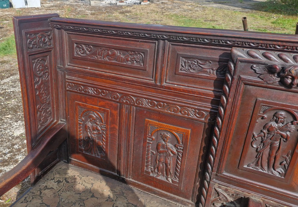 A dark carved oak hall bench featuring relief-carved figure panels on the high back, padded upholstered seats, and a central drop-down table section. View XII