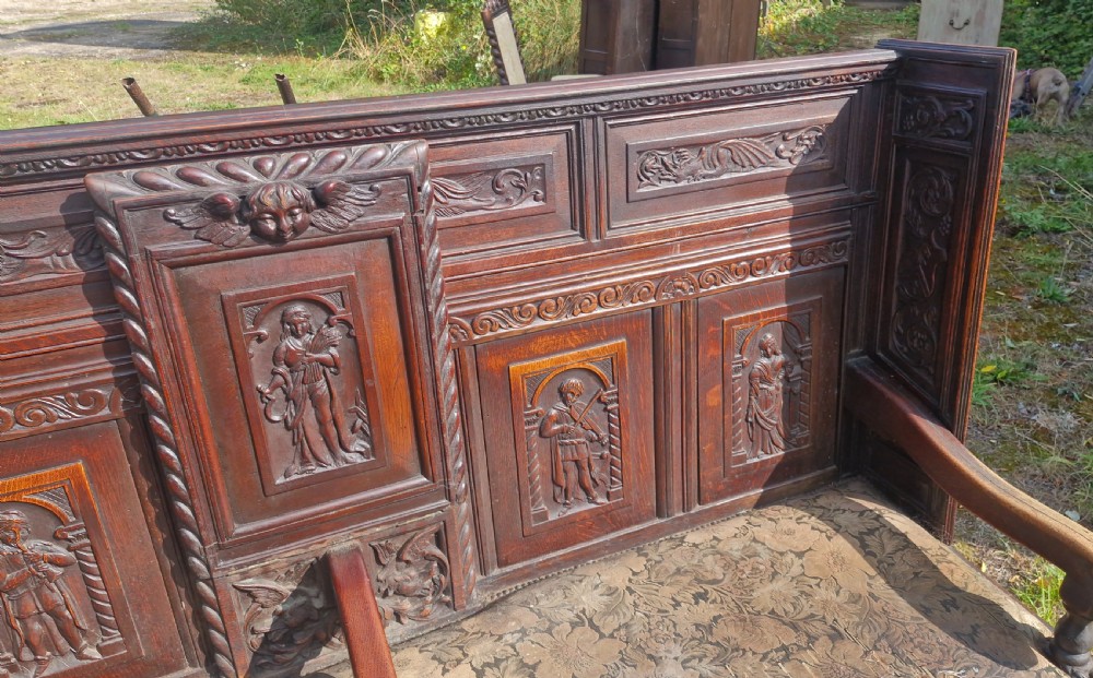 A dark carved oak hall bench featuring relief-carved figure panels on the high back, padded upholstered seats, and a central drop-down table section. View XIII