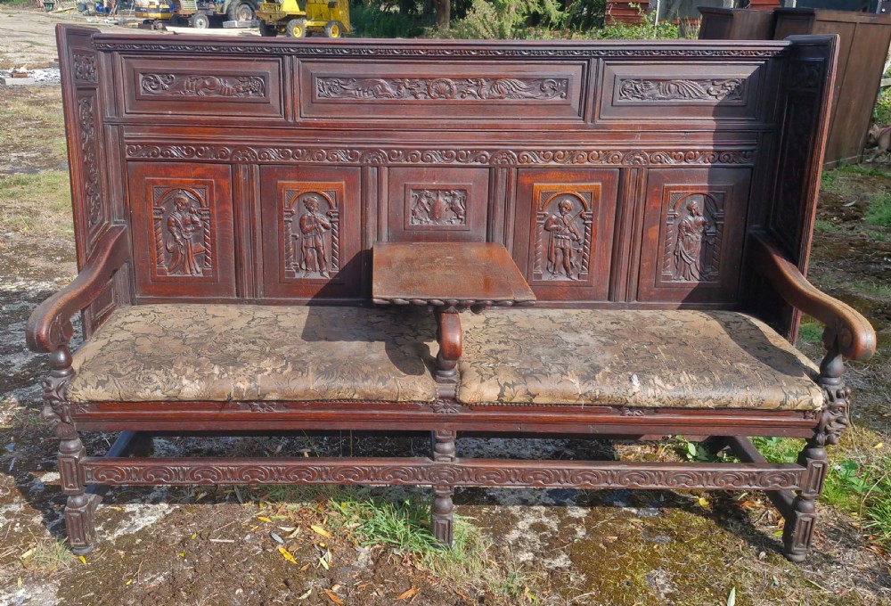 A dark carved oak hall bench featuring relief-carved figure panels on the high back, padded upholstered seats, and a central drop-down table section. View XIV