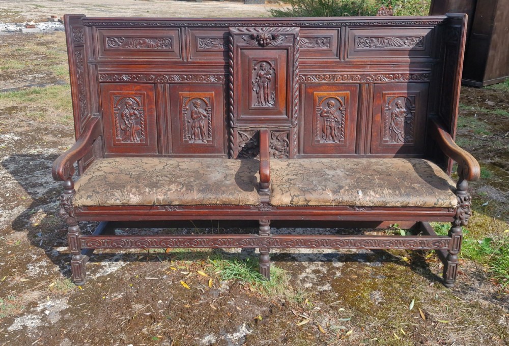 A dark carved oak hall bench featuring relief-carved figure panels on the high back, padded upholstered seats, and a central drop-down table section. View XVI