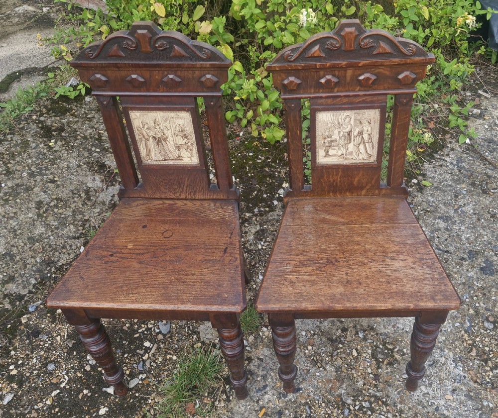 A pair of dark oak hall chairs featuring inset ceramic tiles with historic figures in the backrests, solid seats, and turned front legs.