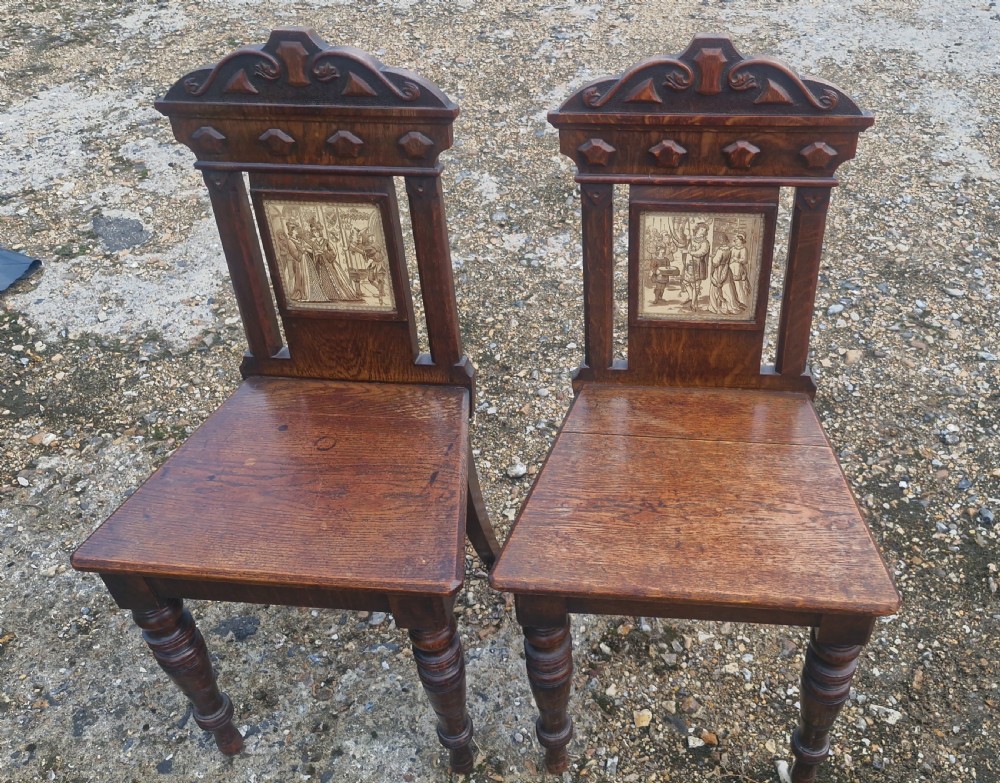 A pair of dark oak hall chairs featuring inset ceramic tiles with historic figures in the backrests, solid seats, and turned front legs. View IV