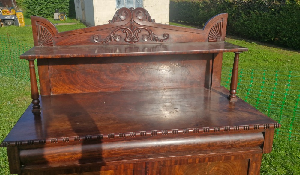 A Regency mahogany two-door side cabinet featuring a raised shelf with turned posts, carved cresting, and panelled flame mahogany doors. View VI