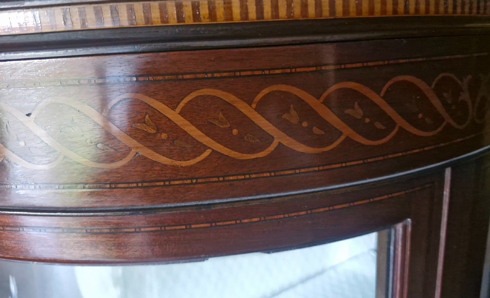 An Edwardian demi-lune mahogany display cabinet with curved glass sides, inlaid decorative banding along the top, and light interior shelves raised on slender legs. View II