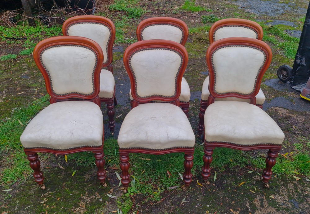 A set of six mid-Victorian mahogany dining chairs with cream upholstered balloon backs, brass studding, and turned front legs on castors. View X