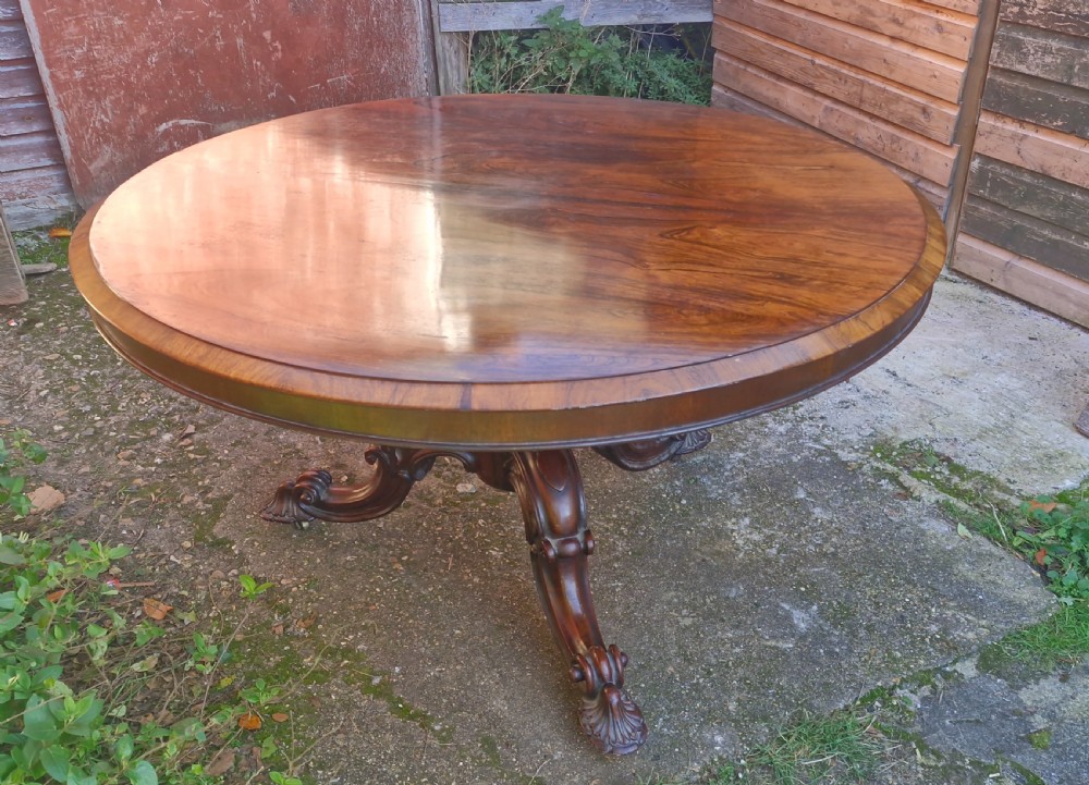 A circular mid-19th-century rosewood centre table featuring a polished top, deep apron, and ornate carved pedestal legs. View VII