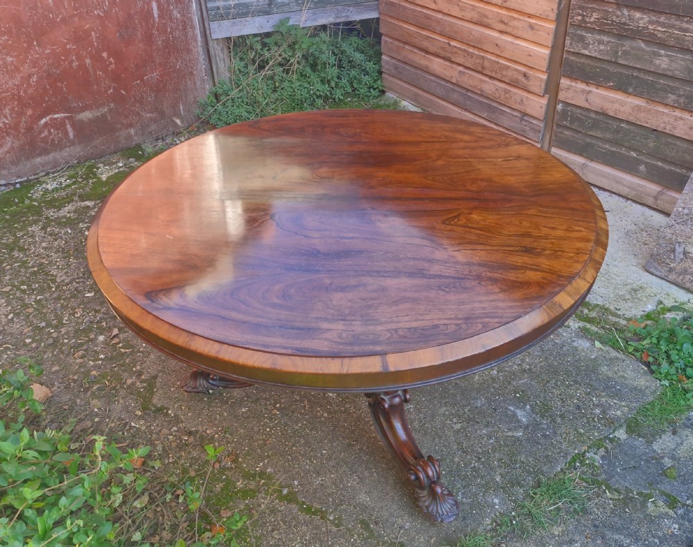 A circular mid-19th-century rosewood centre table featuring a polished top, deep apron, and ornate carved pedestal legs. View IX