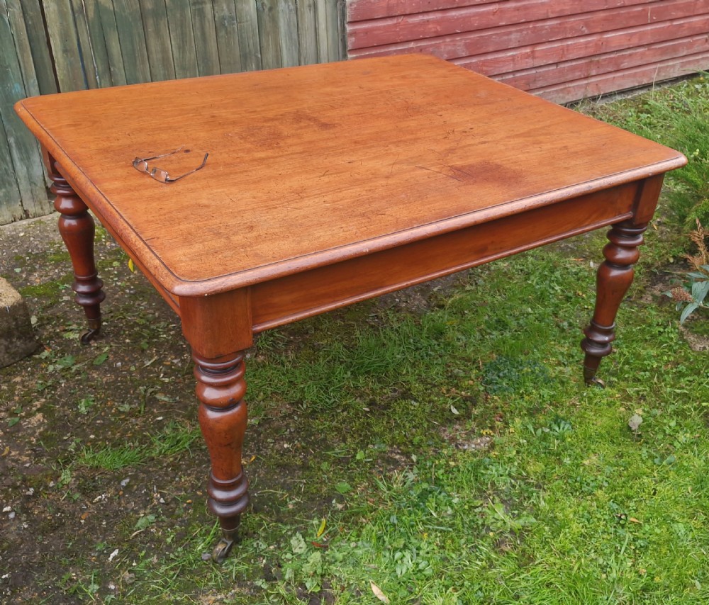 A square Victorian wooden kitchen dining table featuring turned legs fitted with brass castors.