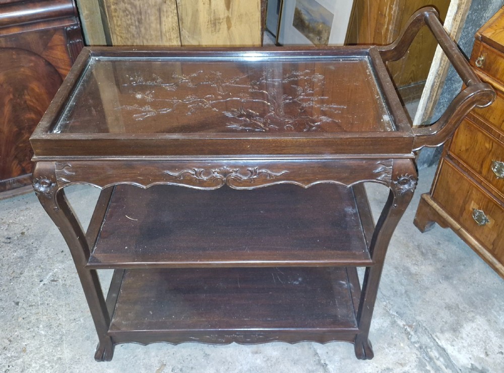 A dark hardwood three-tier tea trolley featuring a carved floral panel under a glass top insert, raised on carved legs with an integrated handle. View II