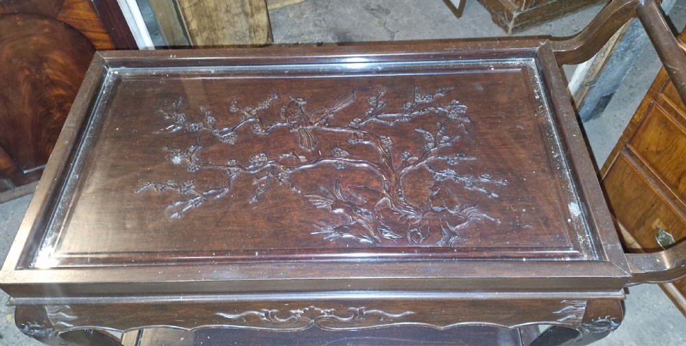 A dark hardwood three-tier tea trolley featuring a carved floral panel under a glass top insert, raised on carved legs with an integrated handle. View III