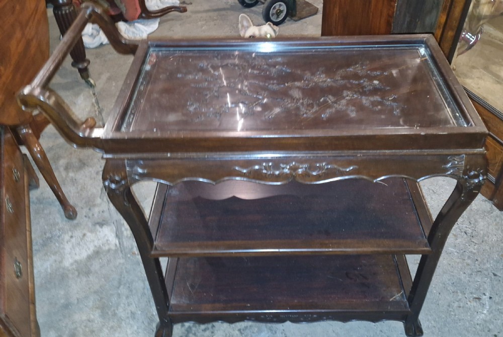 A dark hardwood three-tier tea trolley featuring a carved floral panel under a glass top insert, raised on carved legs with an integrated handle. View V