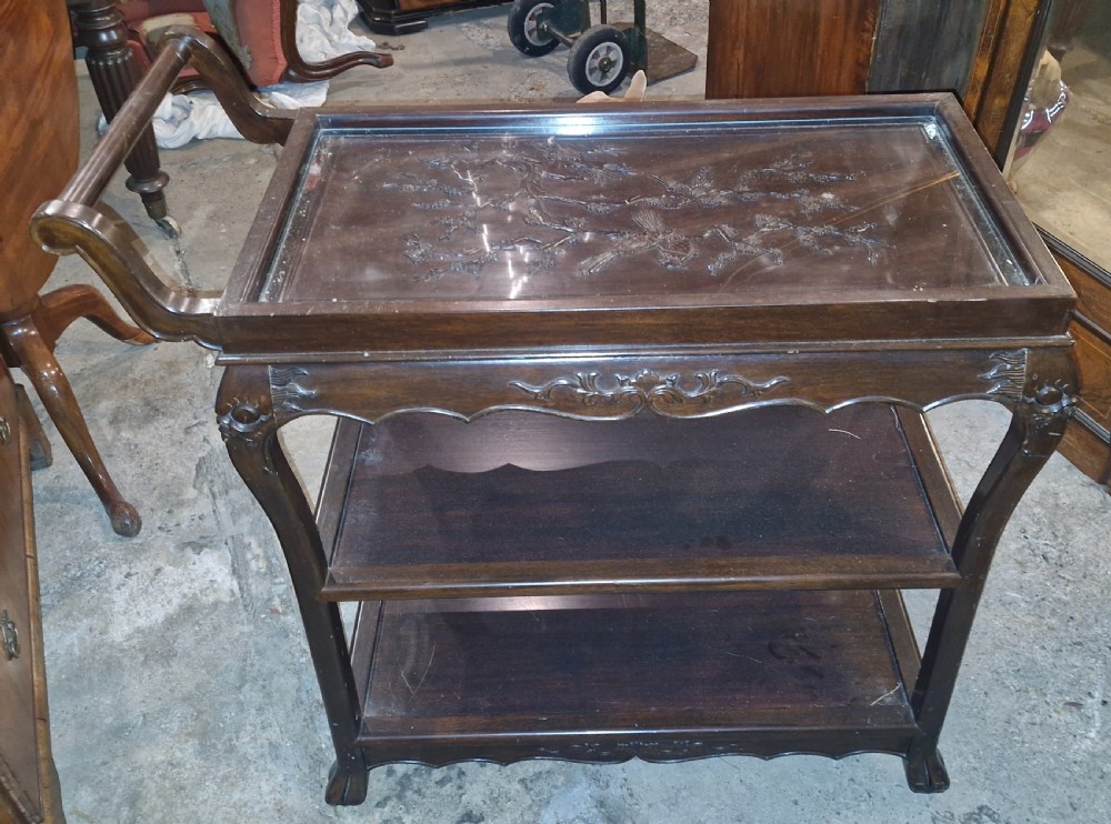A dark hardwood three-tier tea trolley featuring a carved floral panel under a glass top insert, raised on carved legs with an integrated handle. View VI