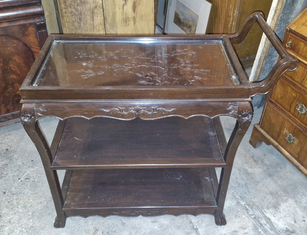 A dark hardwood three-tier tea trolley featuring a carved floral panel under a glass top insert, raised on carved legs with an integrated handle.