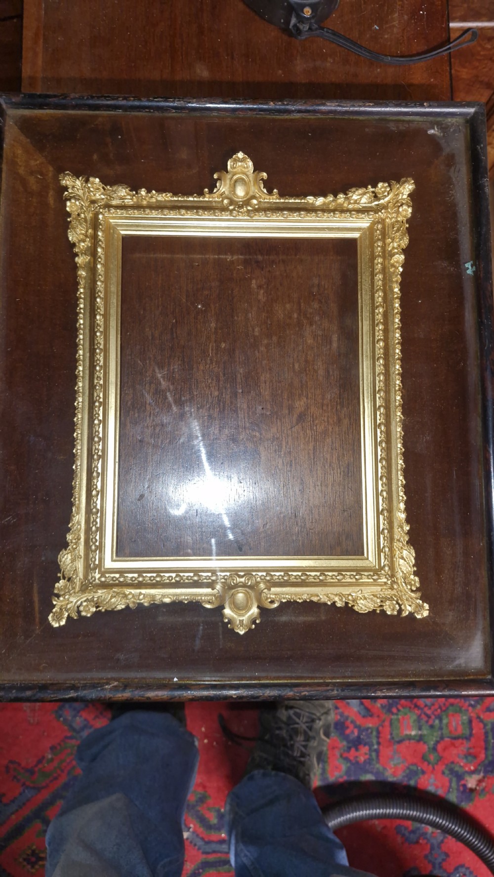 An ornate rectangular gilt picture frame with scrolling foliage cresting, set inside a dark wooden frame. View IV