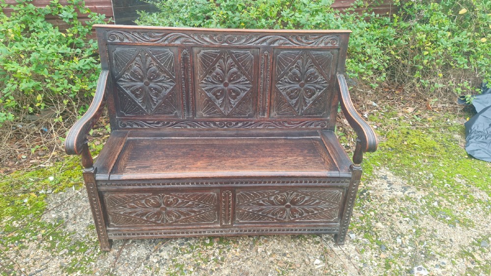 A dark carved oak hall bench featuring three relief-carved back panels, shaped armrests, and a hinged seat opening to a storage chest.