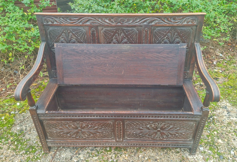 A dark carved oak hall bench featuring three relief-carved back panels, shaped armrests, and a hinged seat opening to a storage chest. View II