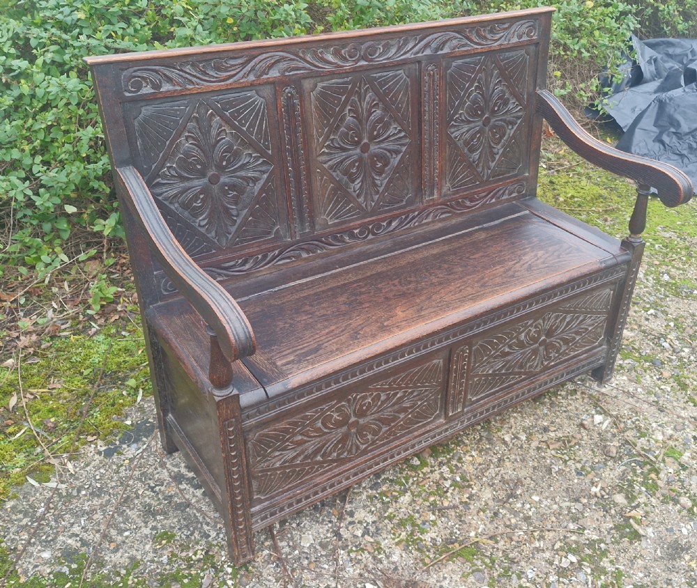 A dark carved oak hall bench featuring three relief-carved back panels, shaped armrests, and a hinged seat opening to a storage chest. View III