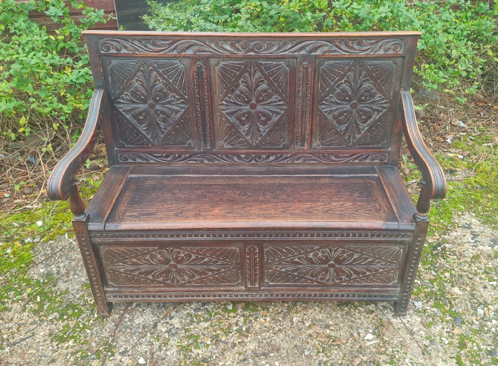 A dark carved oak hall bench featuring three relief-carved back panels, shaped armrests, and a hinged seat opening to a storage chest. View V