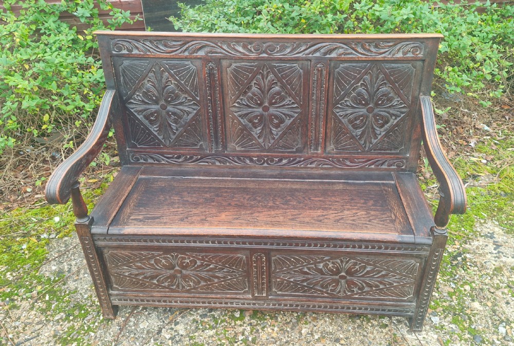 A dark carved oak hall bench featuring three relief-carved back panels, shaped armrests, and a hinged seat opening to a storage chest. View VI