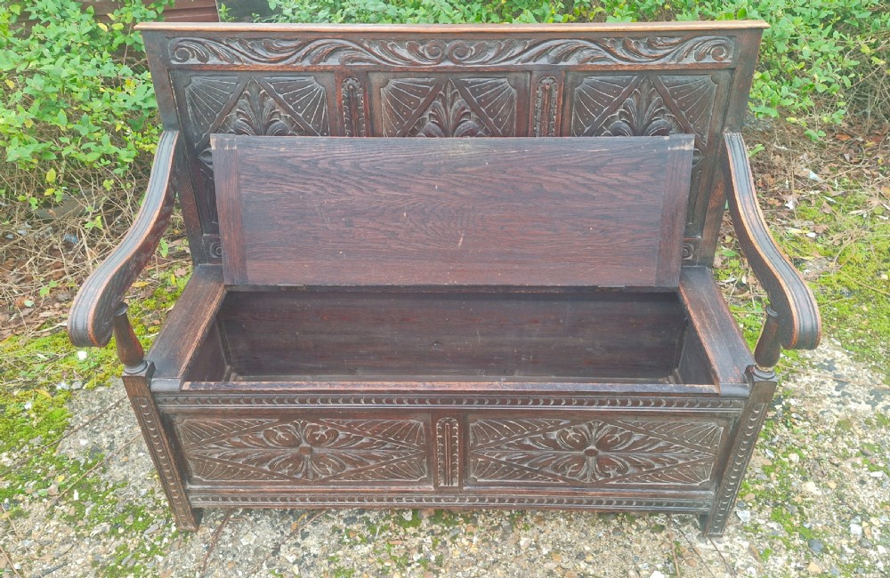 A dark carved oak hall bench featuring three relief-carved back panels, shaped armrests, and a hinged seat opening to a storage chest. View VII