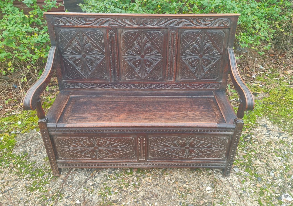A dark carved oak hall bench featuring three relief-carved back panels, shaped armrests, and a hinged seat opening to a storage chest. View VIII