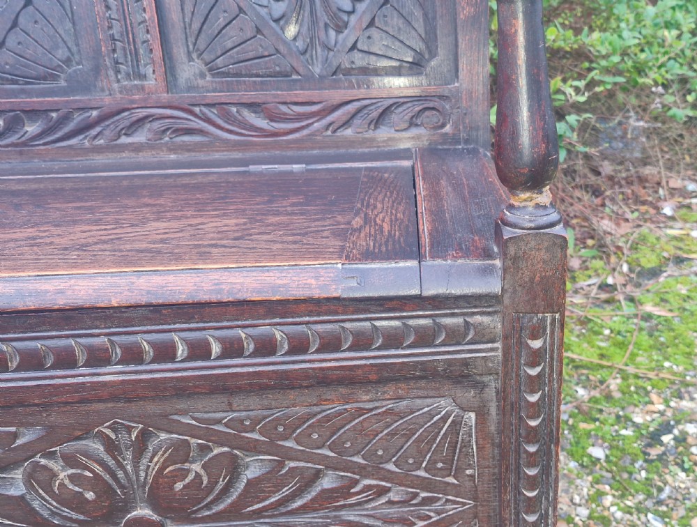 A dark carved oak hall bench featuring three relief-carved back panels, shaped armrests, and a hinged seat opening to a storage chest. View IX