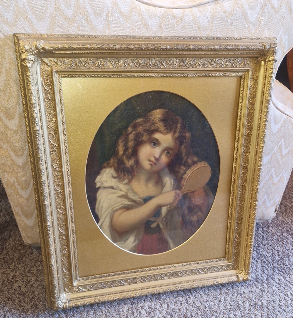 Late Victorian oil painting on canvas depicting a young girl brushing her brown hair, framed in an ornate rectangular gilt frame with an oval spandrel aperture.