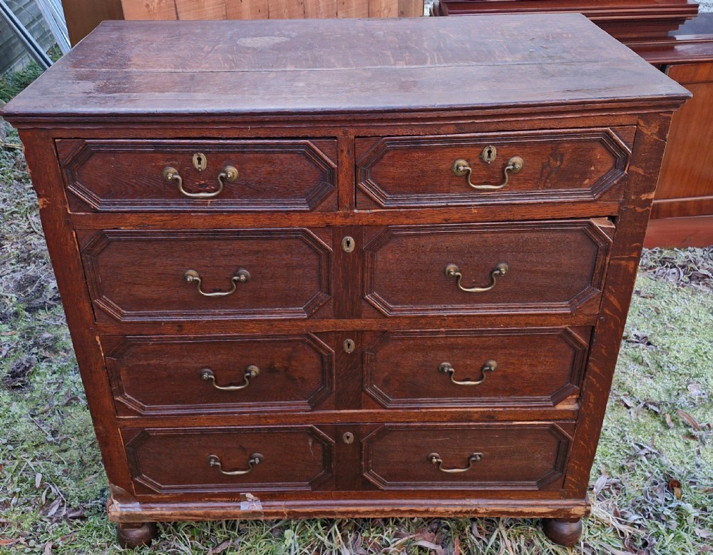 An 18th-century dark oak chest of drawers featuring geometric octagonal panel mouldings, brass drop handles, keyhole escutcheons, and turned bun feet.