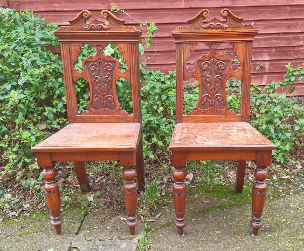 A pair of late Victorian wooden hall chairs featuring ornate carved back splats, crest pediments, solid wood seats, and turned front legs.