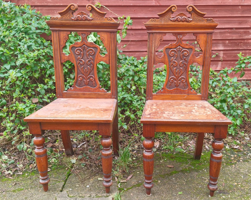 A pair of late Victorian wooden hall chairs featuring ornate carved back splats, crest pediments, solid wood seats, and turned front legs. View II