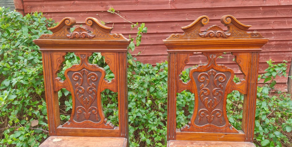 A pair of late Victorian wooden hall chairs featuring ornate carved back splats, crest pediments, solid wood seats, and turned front legs. View III