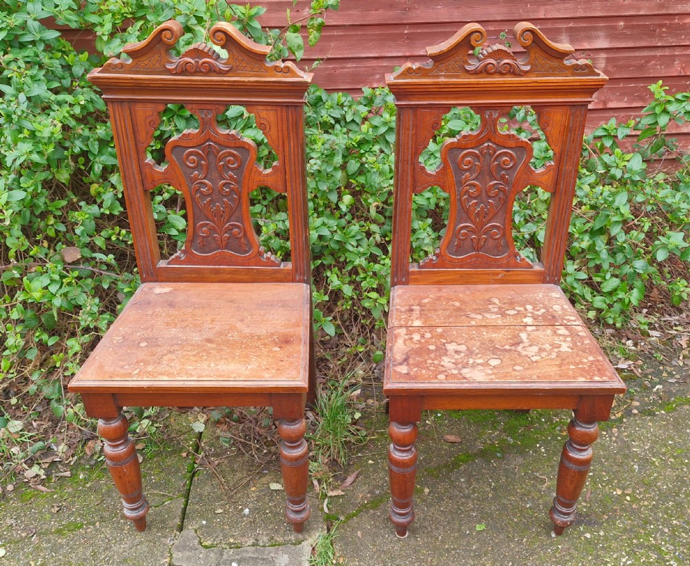 A pair of late Victorian wooden hall chairs featuring ornate carved back splats, crest pediments, solid wood seats, and turned front legs. View V