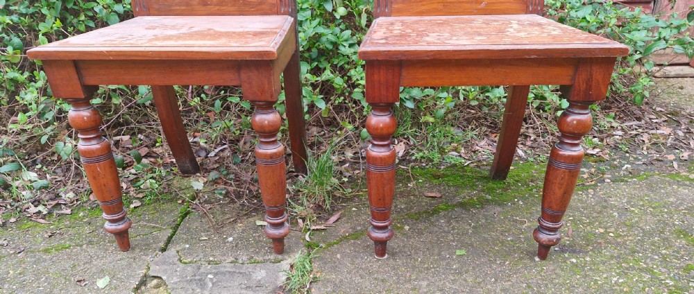 A pair of late Victorian wooden hall chairs featuring ornate carved back splats, crest pediments, solid wood seats, and turned front legs. View VI