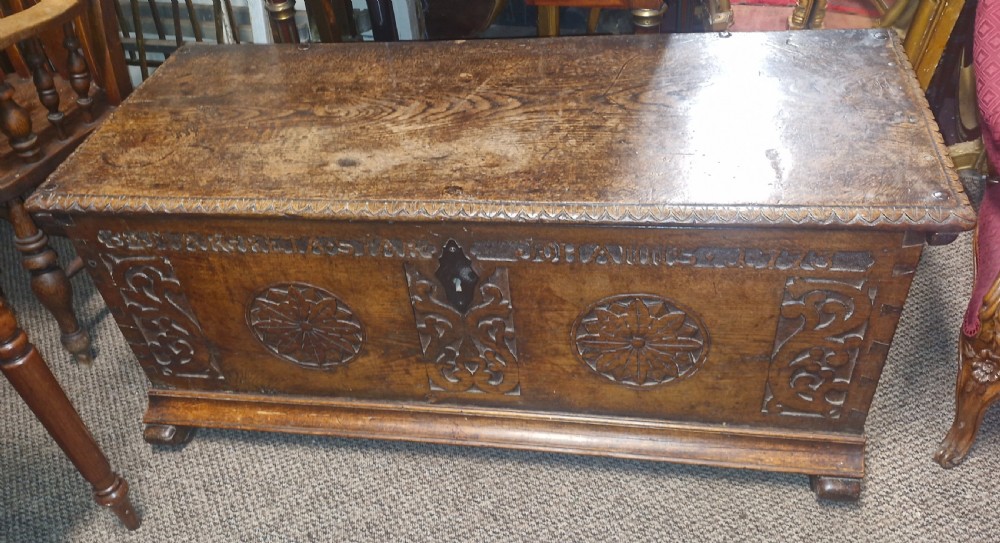 A dark brown 18th-century carved oak blanket chest featuring rosette relief carvings, a carved top frieze, and a central iron escutcheon. View V