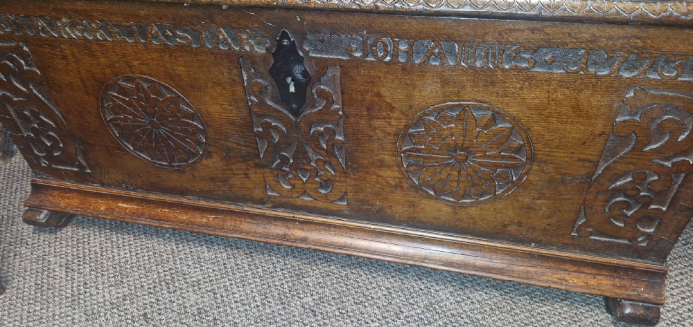 A dark brown 18th-century carved oak blanket chest featuring rosette relief carvings, a carved top frieze, and a central iron escutcheon. View XVIII