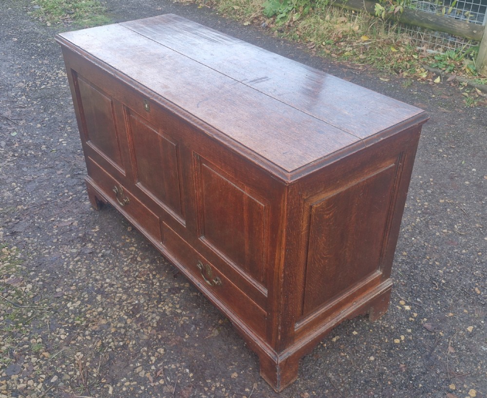 A late Georgian oak blanket chest featuring three fielded front panels, two lower drawers with brass drop handles, and a hinged lid. View V